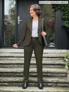 Miik founder Donna (5'6", small) smiling and looking away wearing Miik's Emily soft blazer in olive, regular fit, with a dress pant in the same colour, a black belt and a newport stripe tee, boots and a black purse #color_olive 