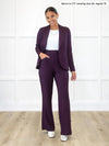 Miik model Meron (5'3", xsmall) smiling while standing in front of a white wall wearing Miik's Emily soft blazer in port, regular fit, a wide leg pant in the same colour and a white tank #color_port 