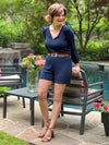 Woman leaning on the back of a garden chair with a pool in the background, smiling and looking away while wearing Miik's Janice 3/4 sleeve short romper in navy with a brown belt and sandals. #color_navy