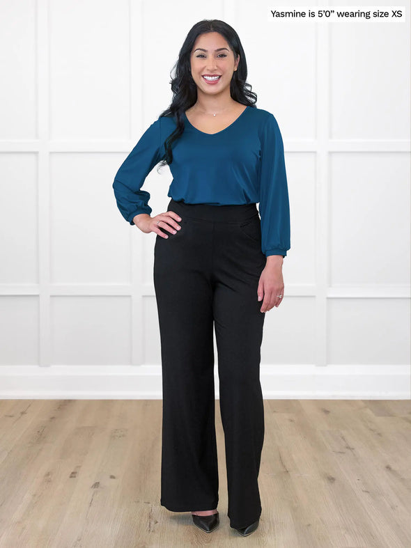 Miik model Yasmine (5'0", xsmall, petite) smiling while standing in front of a white wall wearing a wide leg black pant along with Miik's Jayna v-neck 3/4 sleeve top in teal #color_teal  