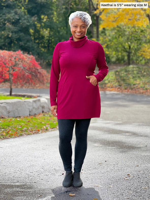 Miik model Keethai (5'5", medium) smiling with one hand on pocket wearing Miik's Kaito cowl neck pocket tunic in cerise with a black legging #color_cerise 