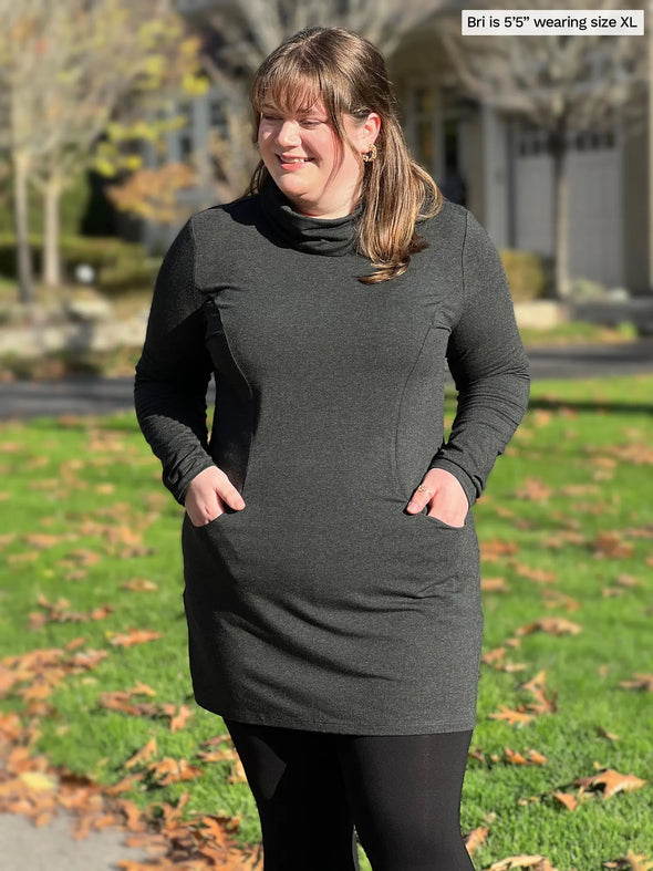 Miik model Bri (5'5", xlarge) smiling and looking down wearing Miik's Kaito cowl neck pocket tunic in charcoal with leggings #color_charcoal 