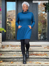 Miik model Keethai (5'5",  medium) smiling wearing Miik's Kaito cowl neck pocket tunic in teal with black leggings and boots