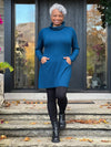 Miik model Keethai (5'5",  medium) smiling wearing Miik's Kaito cowl neck pocket tunic in teal with black leggings and boots #color_teal