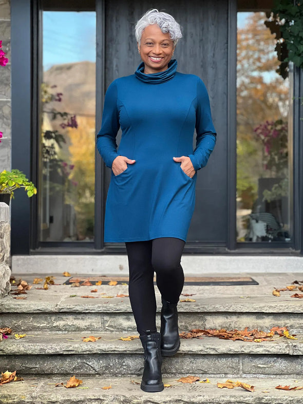 Miik model Keethai (5'5",  medium) smiling wearing Miik's Kaito cowl neck pocket tunic in teal with black leggings and boots #color_teal