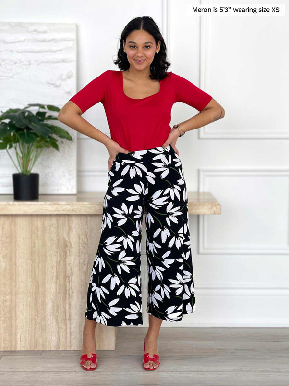 Woman standing in the living room wearing Miik's Keethai wide leg culotte in white floral print with a red top.
#color_white-lily