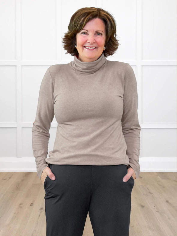 Miik founder Donna (5'6", small) smiling while standing in front of a white wall wearing Miik's Kerry turtleneck top in mushroom melange, regular fit, with a charcoal jogger pant 