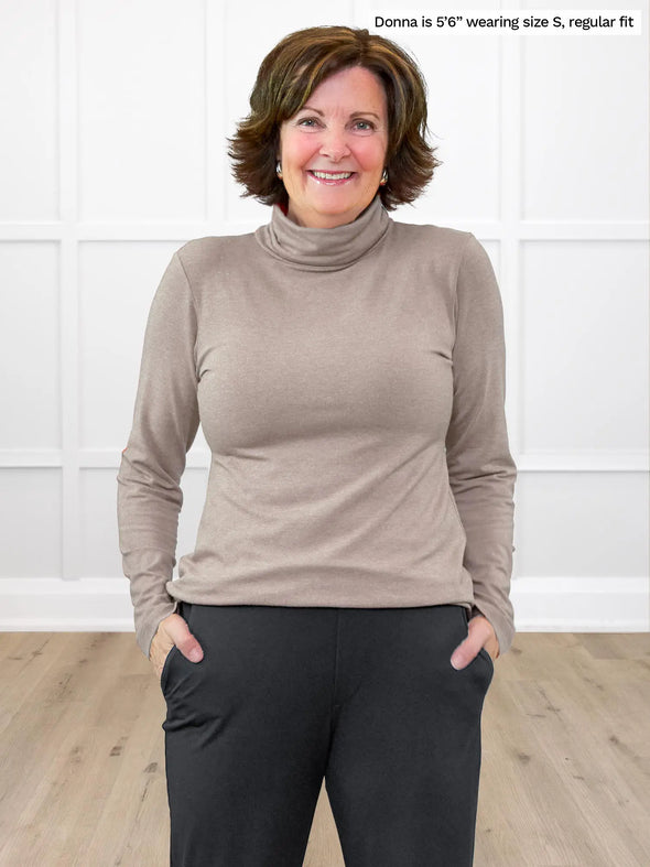 Miik founder Donna (5'6", small) smiling while standing in front of a white wall wearing Miik's Kerry turtleneck top in mushroom melange, regular fit, with a charcoal jogger pant #color_mushroom-melange