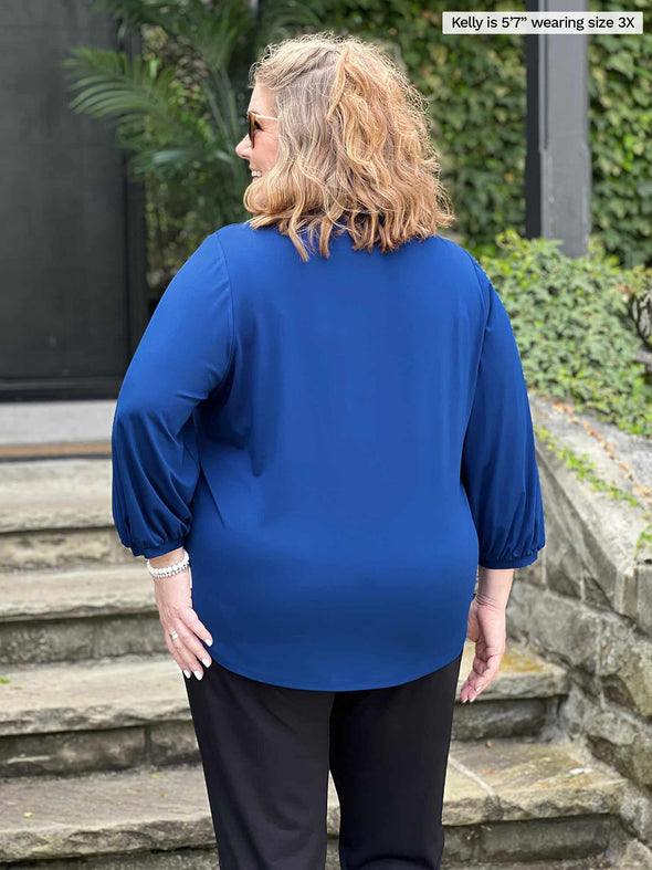 Miik model plus size Kelly (5’7”, 3x) standing with her back towards the camera showing the back of Miik's Kimesha v-neck puff sleeve blouse in ink blue 