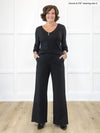 Miik founder Donna (5'6", small) smiling while standing in front of a white wall wearing an all black outfit: Miik's Kiva high waisted wide leg ponte pant with pockets and a v-neck long sleeve blouse #color_black