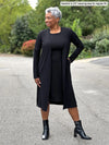 Miik model Keethai (5'5", medium) smiling and looking away wearing Miik's Larissa duster cardigan with pockets in black, regular fit, with a matching colour dress and boots #color_black