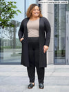 Miik model Carley (5'2", xxlarge) smiling wearing Miik's Larissa duster cardigan with pockets in black, petite fit, with a black dress pant and a mushroom melange tee #color_black