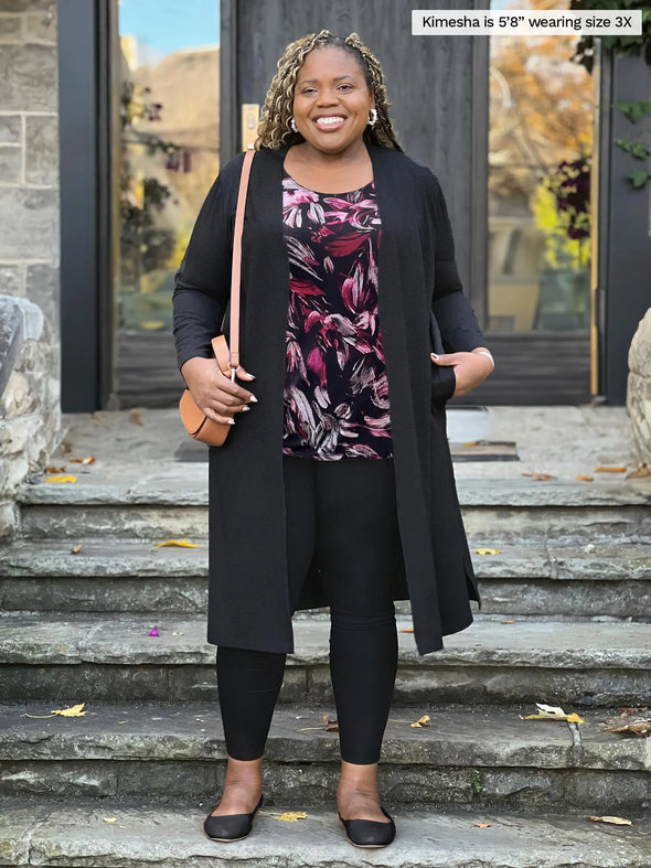 Miik model Kimesha (5'8", 3x) smiling wearing Miik's Larissa duster cardigan with pockets in black with a legging in the same colour and a midnight floral print tank #color_black 