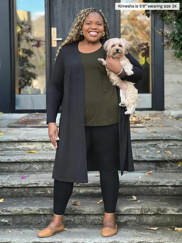 Miik model Kimesha (5'8", 3x) smiling while holding a puppy wearing Miik's Larissa duster cardigan with pockets in charcoal with a black legging and a olive tank top #color_charcoal 