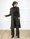 Miik founder Donna (5'6", small) smiling while standing in front of a white wall and looking away wearing Miik's Larissa duster cardigan with pockets in olive with a wide leg pant in the same matching colour and a turtle neck top in black #color_olive