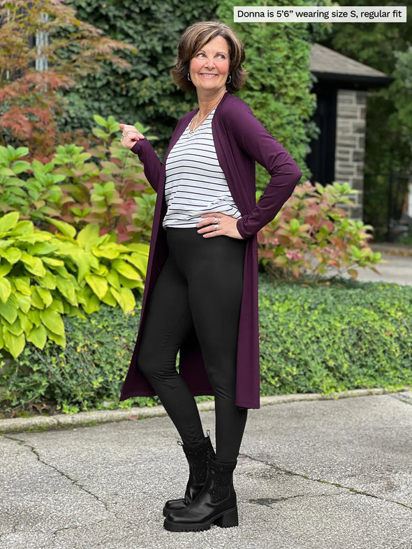 Miik founder Donna (5'6", small) smiling while standing side way 
wearing a black legging, a striped tee, black boots and Miik's Larissa duster cardigan with pockets in port, regular fit  #color_port