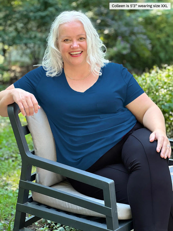 Miik model Colleen (5'3", xxlarge) smiling while sitting on a outdoor chair wearing Miik's Lex v-neck pocket tee in teal and a black dress pant 
#color_teal 