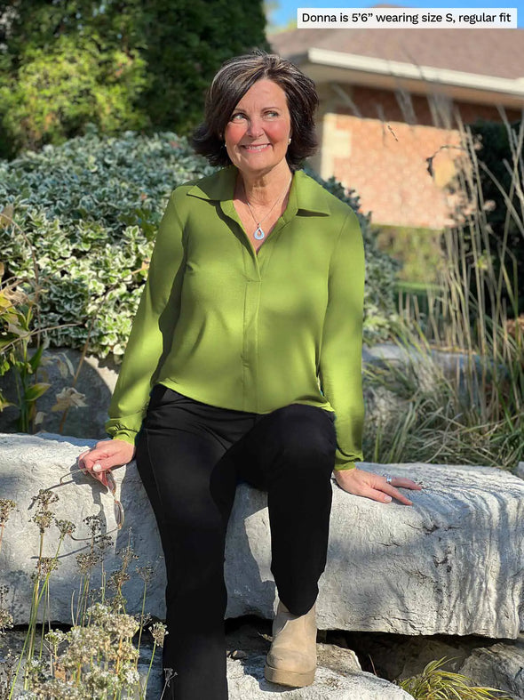 Miik founder Donna (5'6", small) sitting on a rock and looking away wearing Miik's Lucia collared shirt in green moss with a black pant and boots #color_moss