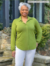 Miik model Keethai (5'5'", medium) smiling wearing Miik's Lucia collared shirt in green moss and a white jeans #color_moss