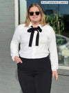 Miik model Bri (5'5", xlarge) smiling while standing in front of a window wearing Miik's Lucia collared shirt in white with a tie bow in black and a black dressy pant #color_white