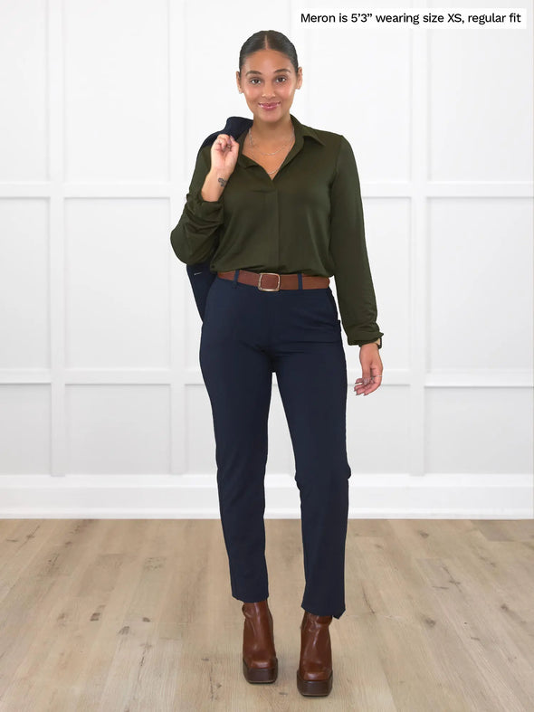 Miik model Meron (5'3", xsmall) smiling while standing in front of a white wall studio wearing Miik's Lucia collared shirt  in olive, regular fit, tucked in a navy pants, brown belt and boots and a blazer over her shoulders 
#color_olive