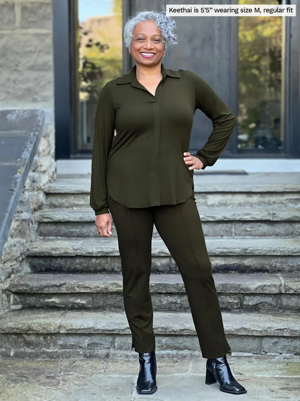 Miik model Keethai (5'5", medium) smiling wearing Miik's Lucia collared shirt in olive, regular fit, along with a dress pant in the same matching colour and black boots 
#color_olive