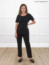 Miik model Lex (5'2", xsmall) smiling while standing in front of a white wall studio wearing Miik's Madeline reversible neckline ribbed half sleeve t-shirt in black with a dress pant in the same pant 
#color_black-ribbed