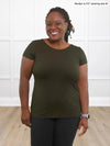 Miik model Marilyn (5'2", medium) smiling while standing in front of a white wall wearing Miik's Marianna reversible classic tee in olive and a charcoal pant 
#color_olive 