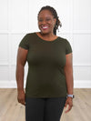 Miik model Marilyn (5'2", medium) smiling while standing in front of a white wall wearing Miik's Marianna reversible classic tee in olive and a charcoal pant 