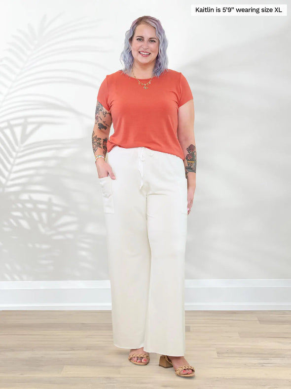 Miik model Kaitlin (5'9", xlarge) smiling while standing in front of a white wall wearing Miik's Marianna reversible neckline classic tee in papaya melange with a wide leg lounge pant in natural #color_papaya-melange