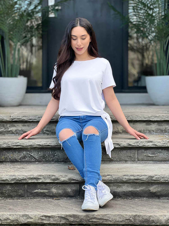 Miik model Yasmine (5'0", xsmall) sitting on a backyard stairs and looking down wearing Miik's Melody side tie top in white with ripped jeans 
#color_white