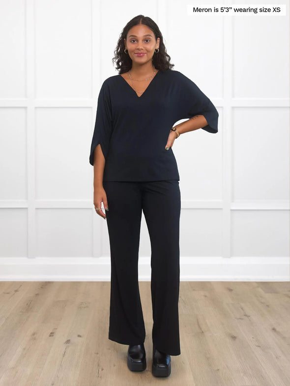 Miik model Meron (5'3", xsmall) smiling while standing in front of a white wall studio wearing Miik's Meron v-neck 3/4 sleeve top in black with a high waisted pant in the same colour
#color_black 