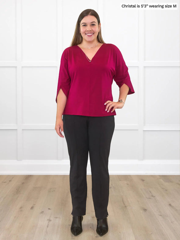 Miik model Christal (5'3", medium) smiling while standing in front of a white wall studio wearing a black dress pant with Miik's Meron v-neck 3/4 sleeve top in cerise and boots 
#color_cerise 