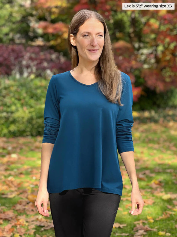 Miik model Lex (5'2", xsmall) smiling wearing Miik's Pippa reversible neckline long sleeve top in teal with a black pant #color_teal 