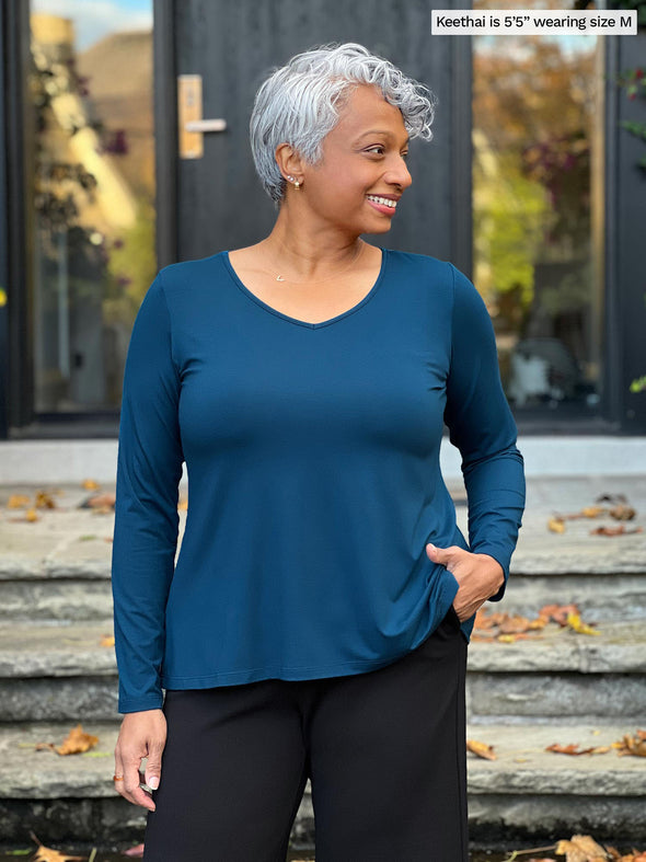 Miik model Keethai (5'5", medium) looking away wearing Miik's Pippa reversible neckline long sleeve top in teal with a black dress pant #color_teal