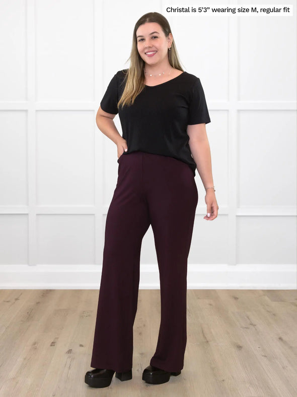 Miik model Christal (5'3", medium) smiling while standing in front of a white wall wearing a black ribbed tee with Miik's Reed high waisted pant with pockets in port, regular fit 
#color_port 