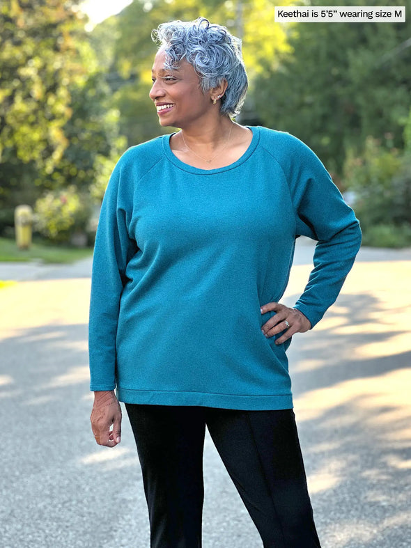 Miik model Keethai (5'5", medium) smiling and looking away wearing a black dress pant with Miik's Ria crewneck lightweight fleece sweatshirt in teal melange 
#color_teal-melange  