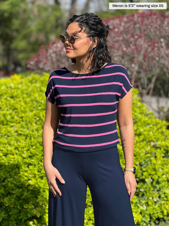 Woman standing in the garden wearing Miik's Rio reversible dolman tee in pink and navy stripe with navy pants.