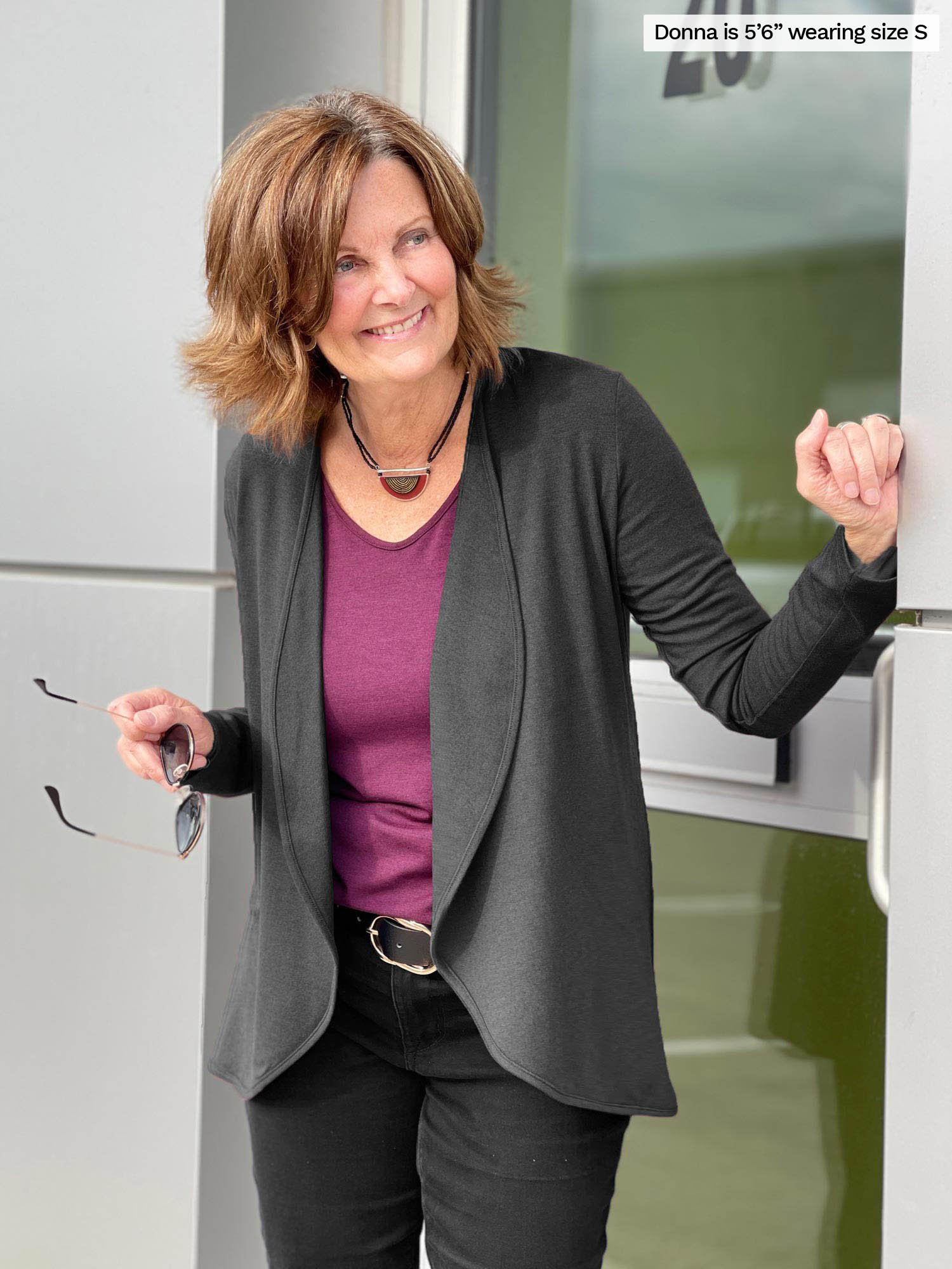 Miik founder Donna (size S, 5 foot 6) wearing the Rory waterfall cardigan in charcoal grey with a port-coloured Shannon top underneath, tucked into belted black jeans.
