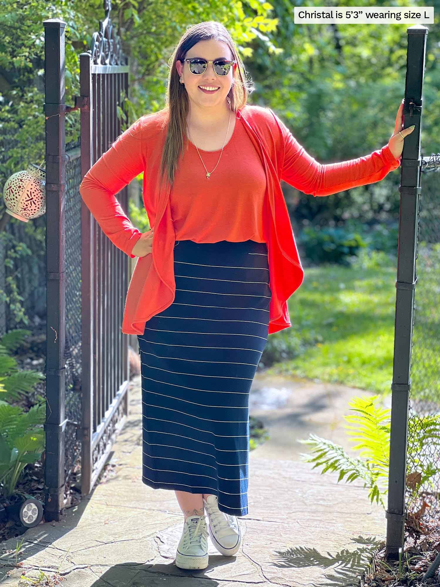 Miik model Christal (five feet three, large) standing next to a backyard gate smiling wearing a pinstripe navy midi skirt along with Miik&