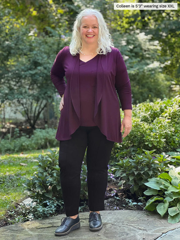 Miik model Colleen (5'3", xxlarge) smiling while standing in front of a garden wearing Miik's Rory waterfall cardigan in port, a tank top in the same colour and a dress pant in black 
#color_port 