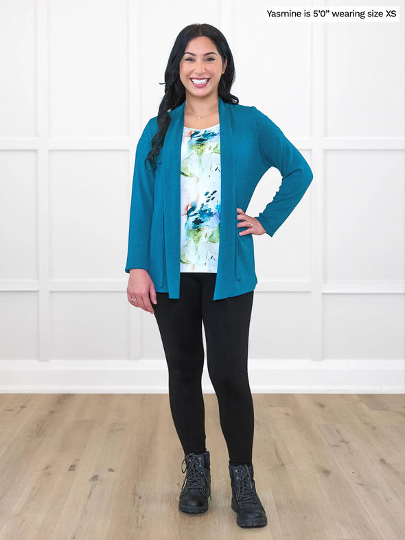 Miik model Yasmine (5'0", xsmall, petite) smiling while standing in front of a white wall wearing a black legging, printed blouse and Miik's Sadira tie waist sweater cardigan in moroccan blue #color_moroccan-blue