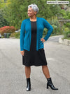 Miik model Keethai (5'5", medium) smiling and looking away wearing a black dress with Miik's Sadira tie waist sweater cardigan in moroccan blue and black boots #color_moroccan-blue 