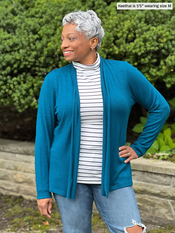 Miik model Keethai (5'5", medium) smiling and looking away wearing Miik's Sadira tie waist sweater cardigan in moroccan blue with a striped turtleneck top and ripped jeans #color_moroccan-blue