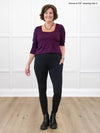 Miik founder Donna (5'6", small) smiling while standing in front of a white wall wearing Miik's Seana high waisted pocket legging in black and a port long sleeve top #color_black