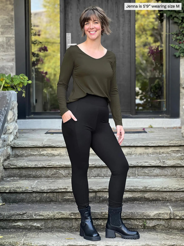 Miik model Jenna (5'9", medium) smiling wearing Miik's Seana high waisted pocket legging in black with a olive long sleeve top and black boots #color_black 