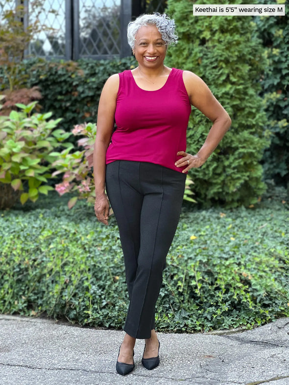 Miik model Keethai (5'5", medium) smiling while standing in front of a garden wearing Miik's Shandra reversible neckline tank top in cerise with a charcoal dress pant 
#color_cerise 