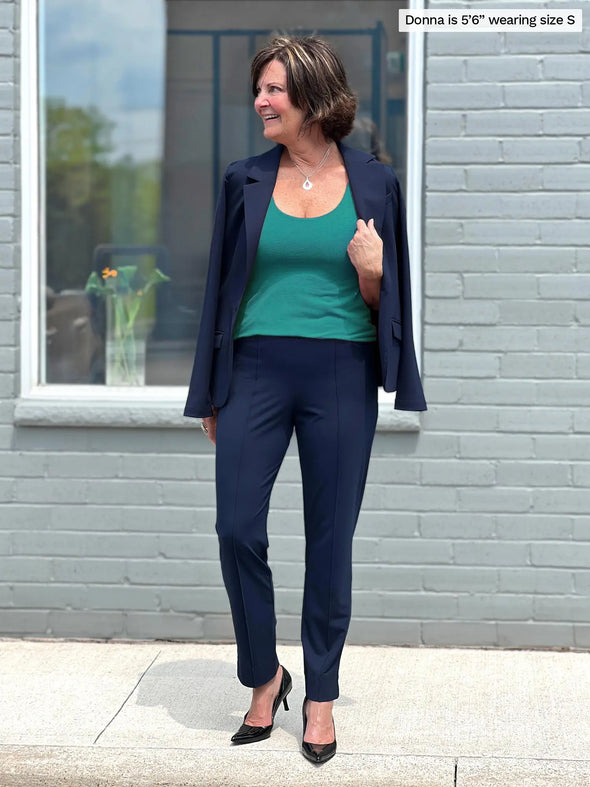 Miik founder Donna (5'6", small) smiling and looking away while standing in front of a brick wall/window wearing Miik's Shandra reversible tank top in jade melange with a navy suit: Christal pant and Emily blazer 
#color_jade-melange 