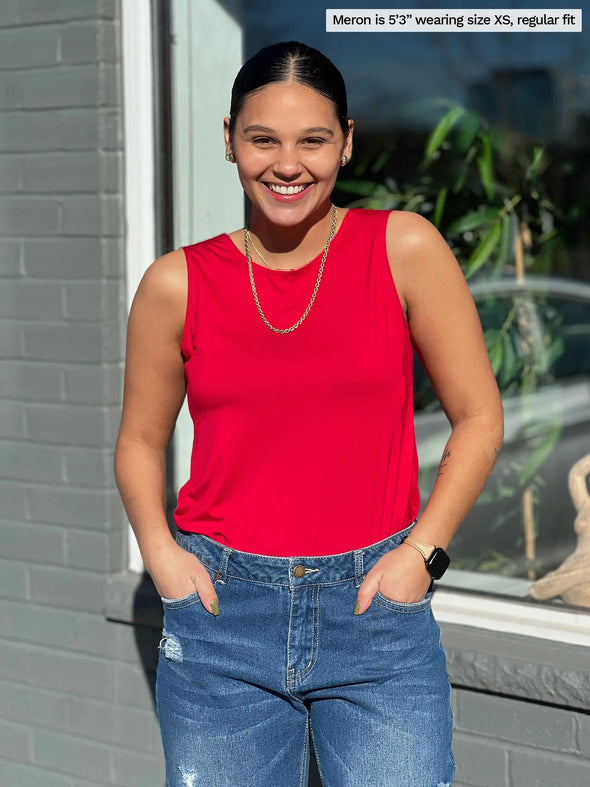 Miik model Meron (5'3", xsmall) smiling while standing in front of a window with both hands on pockets wearing Miik's Shandra reversible tank top in poppy red and jeans
#color_poppy-red 