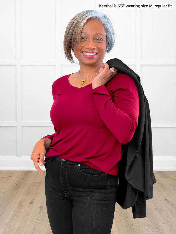 Miik model Keethai (5'5", medium) smiling while standing in front of a white wall wearing Miik's Shannon reversible neckline long sleeve tee in cerise, regula fit, and black jeans #color_cerise 